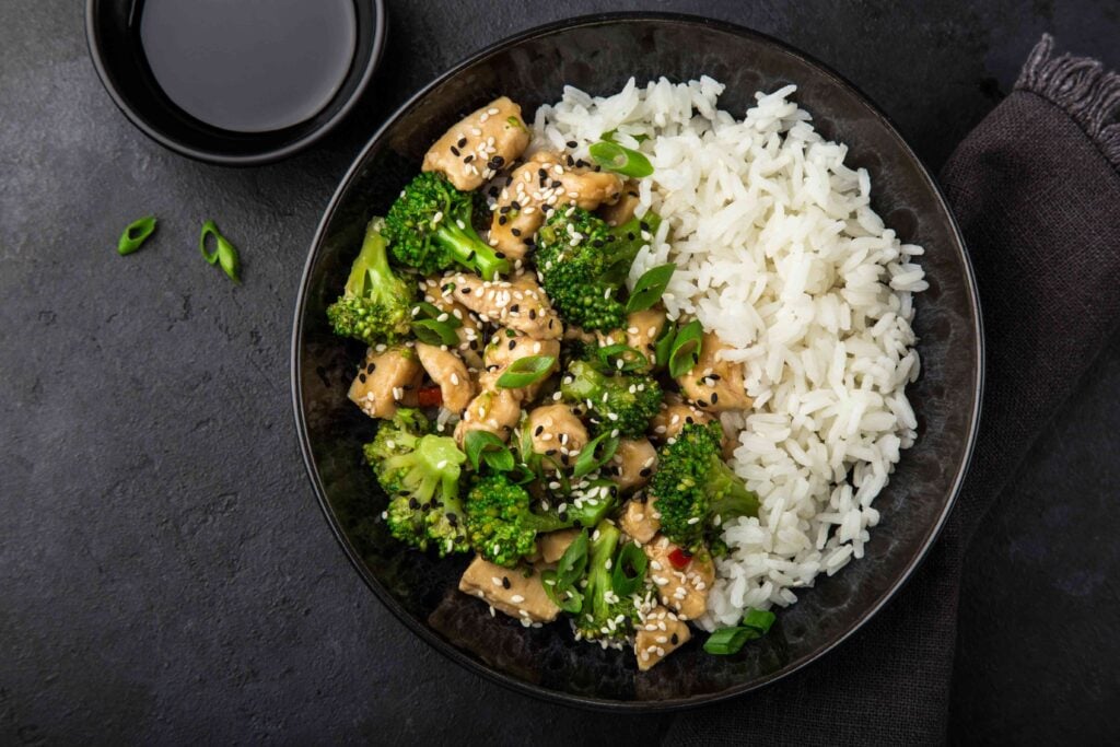Teriyaki chicken and broccoli with steamed rice in bowl, top view, dark background